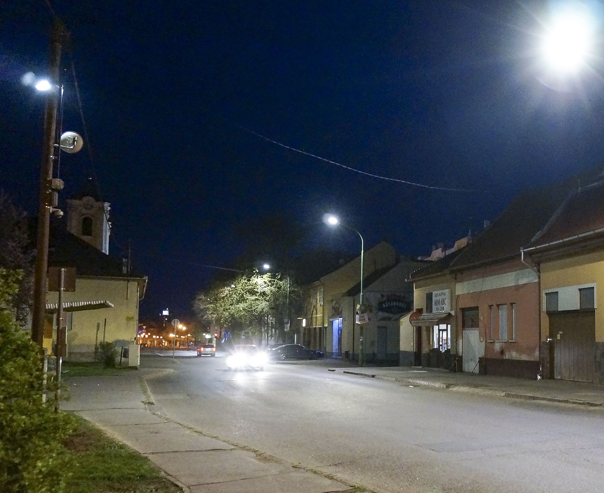 Gyöngyösi utca este, földszintes házakkal, a háttérben templomtorony, az oszlopokon Kombo lámpák.